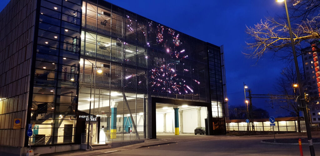Parkeringshus i blåtimmen med rosa ljusburst på fasaden och belysta våningsplan bakom glas.