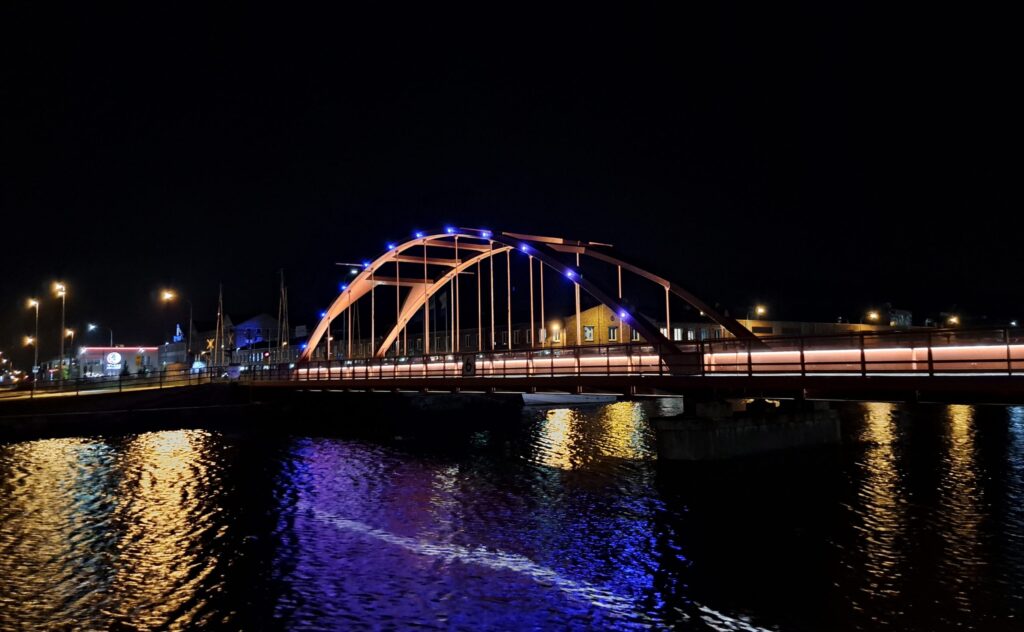 Järnvägsbron i Lidköping nattetid med orange bågar, blå punktljus och ljusstreck från trafik.