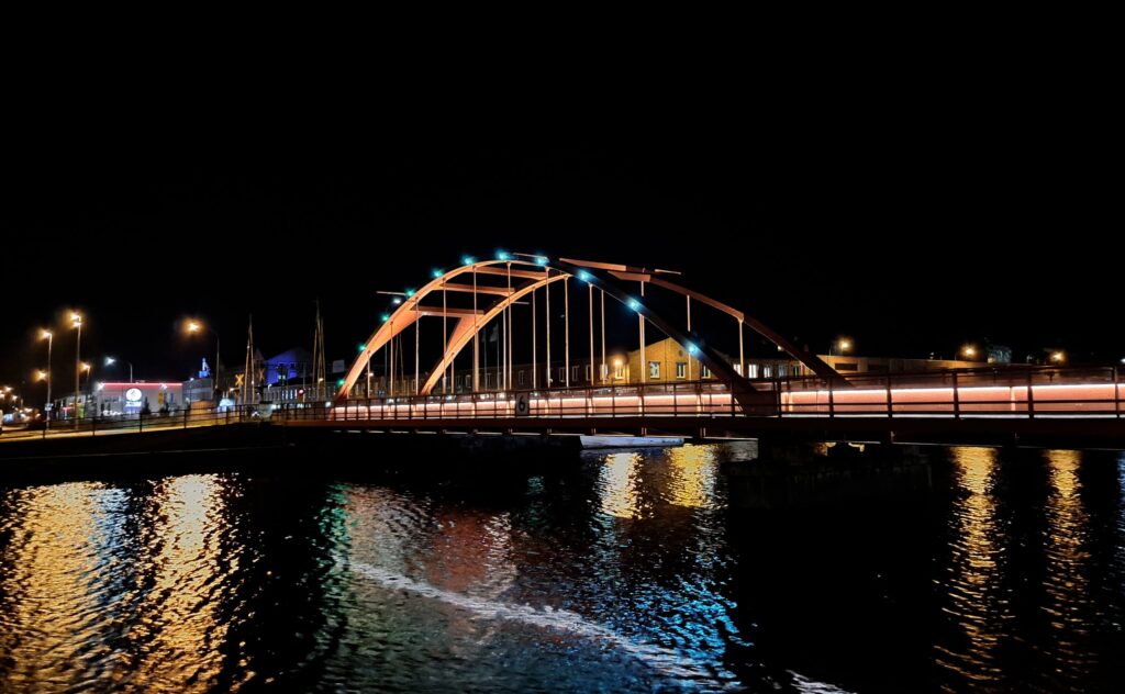 Järnvägsbron i Lidköping nattetid med orange bågar, blå accentljus och reflektion i vattnet.