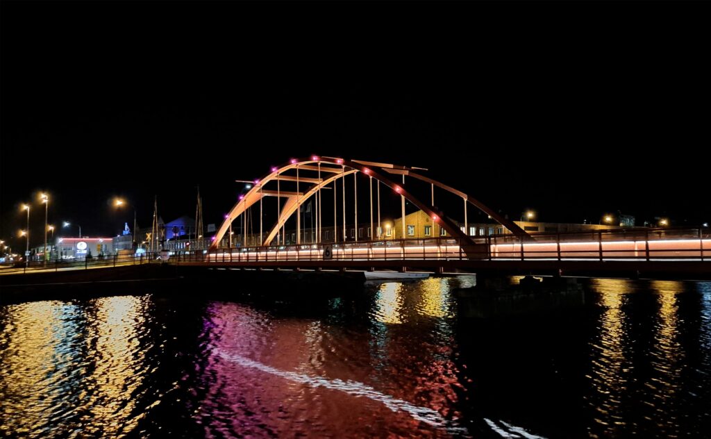 Järnvägsbron i Lidköping nattetid, orange bågbelysning som speglas i sjön mot stadens kaj.