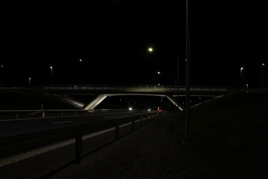 Motorväg i mörker mot belyst överfart, med ljusstreck från fordon under bron.