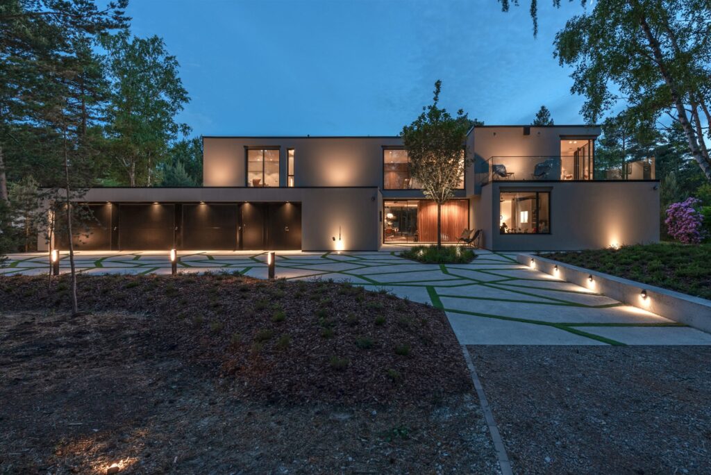 Modern villa i skymning med fasadspotlights, pollare längs infarten och varmt fönsterljus.