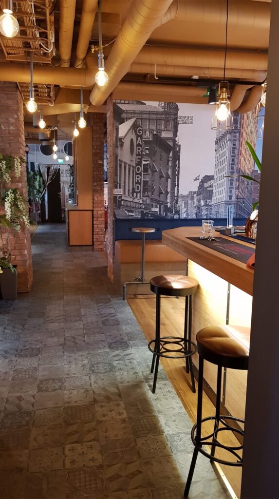 Baren på New York Bar & Grill med hängande glödlampor, exponerade rör och belyst bardisk.