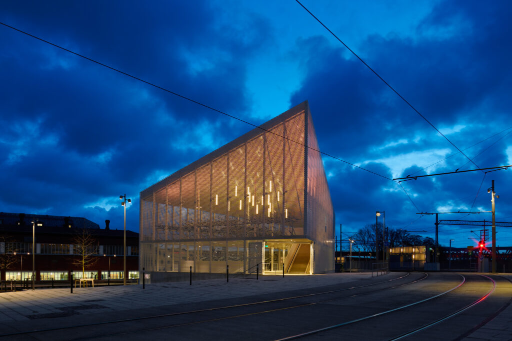 Cykelhuset som en lysande lanternin i guldgult ljus mot indigo himmel och spårvagnsspår.