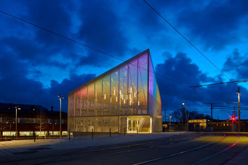 Cykelhuset i blåtimmen med regnbågsskimmer i glasfasaden och vertikala pendellampor inuti.