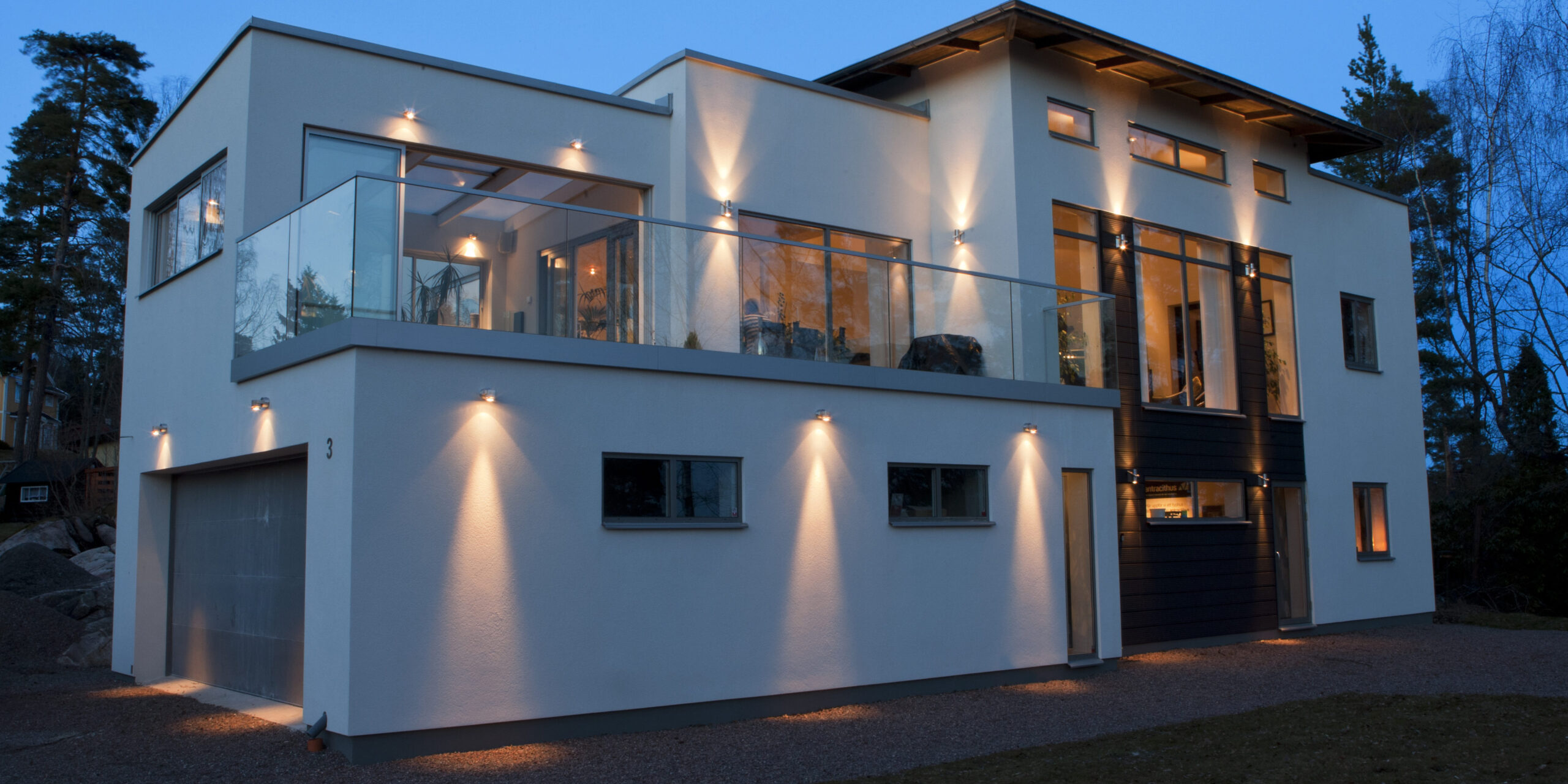 Modern vit villa i skymning med triangulära ljuskäglor från fasadarmaturer och varmt ljus i fönstren.