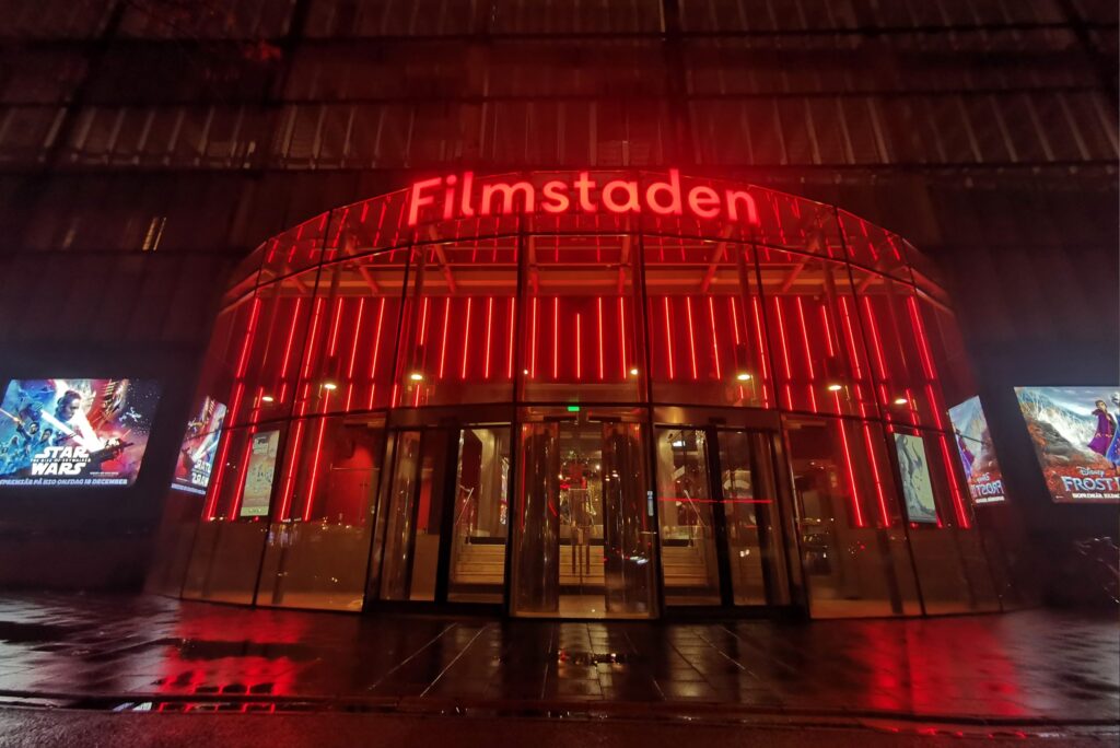 Filmstadens entré på natten med röd neonfasad som speglas i våt asfalt.