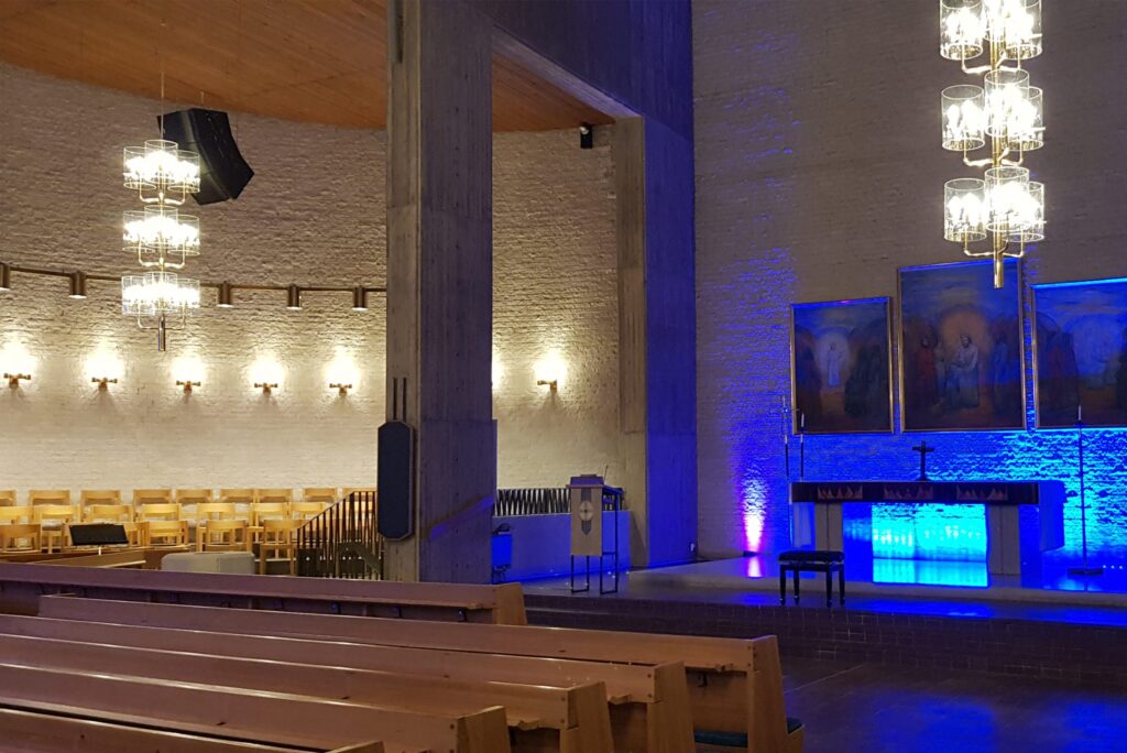 Mikaelikyrkan med blå belyst kor, träbänkar och varma vägglampor längs den putsade väggen.