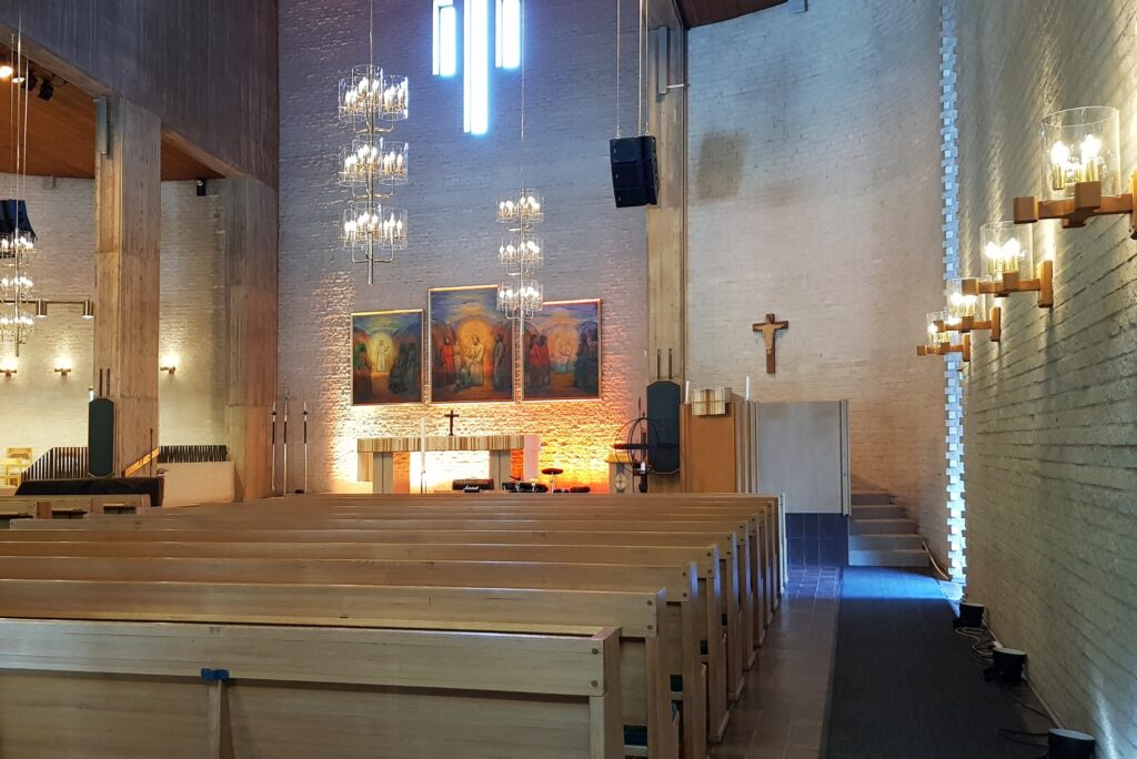 Mikaelikyrkan mot altaret med varm bakgrundsbelysning, triptyk och kristallkronor över träbänkarna.