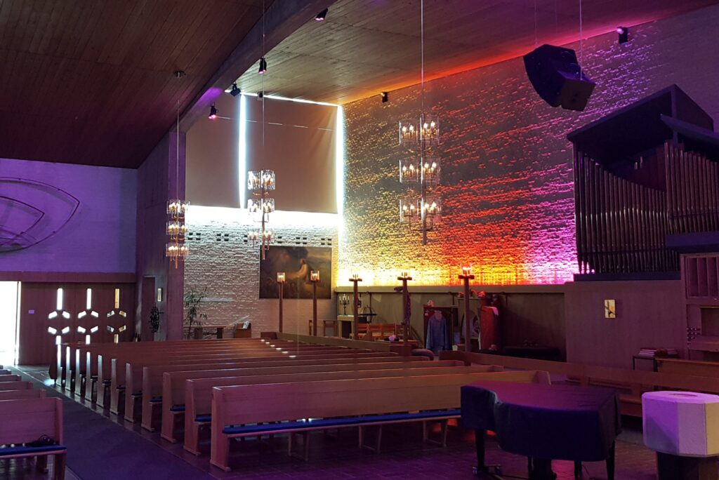 Mikaelikyrkan med lila och orange väggbelysning, orgel till höger och kristallkronor över bänkarna.