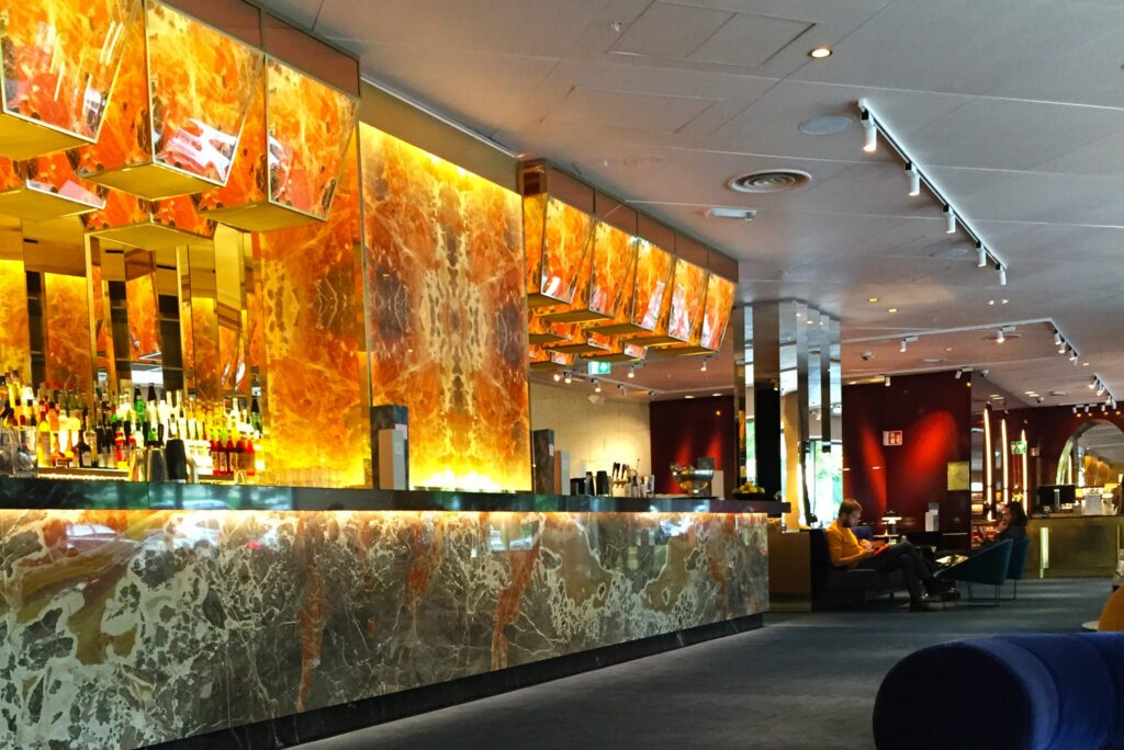 Hotellbar med bakbelyst orange onyxbakstycke, marmorerad bardisk och spotlighttak.