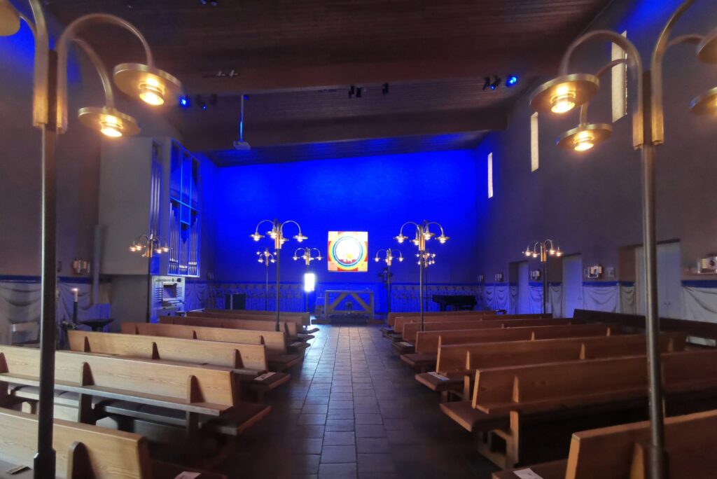 S:t Hans kyrka mot blå korvägg med färgat glasparti över altaret och träbänkar längs stengången.