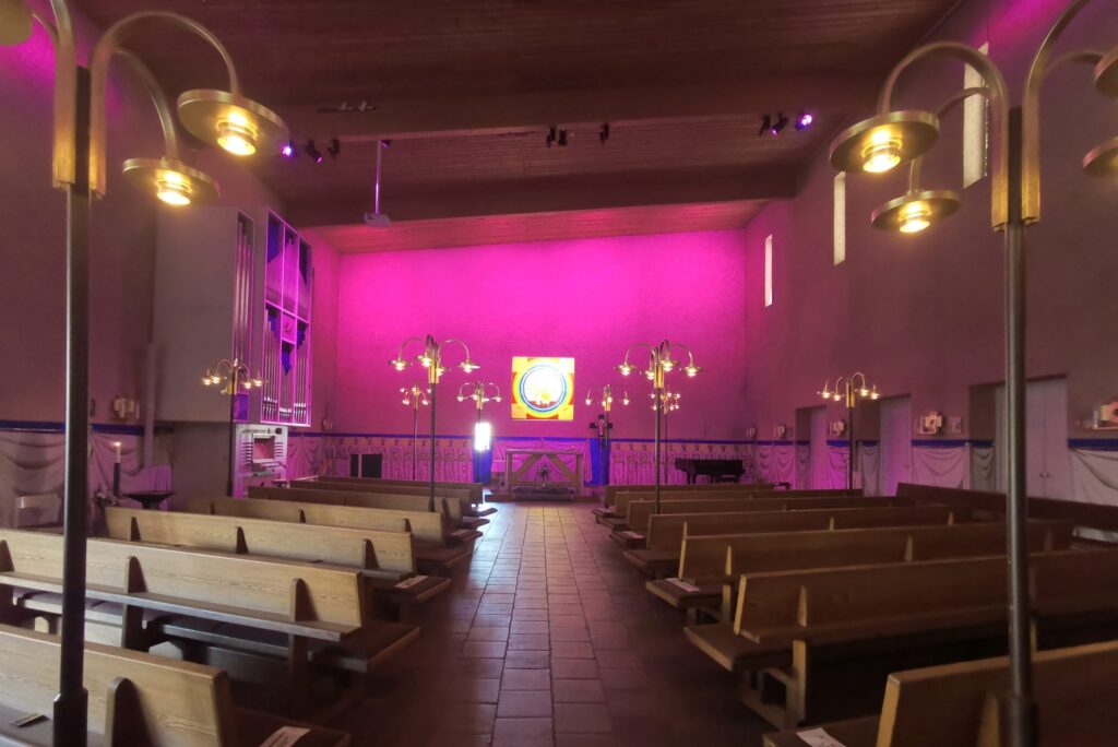 S:t Hans kyrka med magenta belyst korvägg, varmt fönsterljus över altaret och mässingslampor i förgrunden.